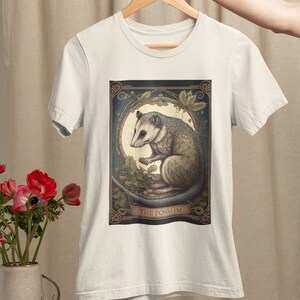 May include: A white t-shirt with a graphic print of a possum in a moonlit forest. The graphic is framed with a decorative border and the text "THE POSSUM" is printed below the image.