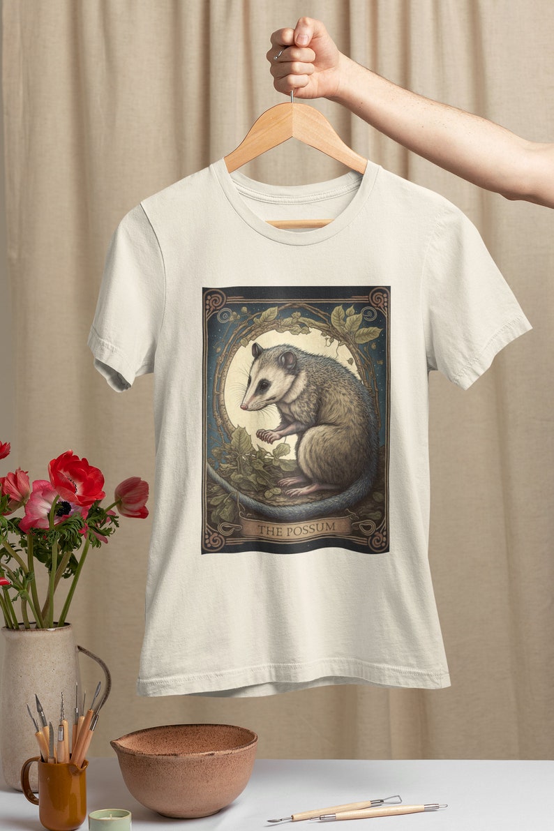 May include: A white t-shirt with a graphic print of a possum in a moonlit forest. The graphic is framed with a decorative border and the text "THE POSSUM" is printed below the image.
