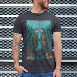 May include: A dark heather grey t-shirt featuring a graphic of two sea otters holding hands in a turquoise underwater scene. The otters are brown with white markings. The scene is surrounded by green and white seaweed and coral.