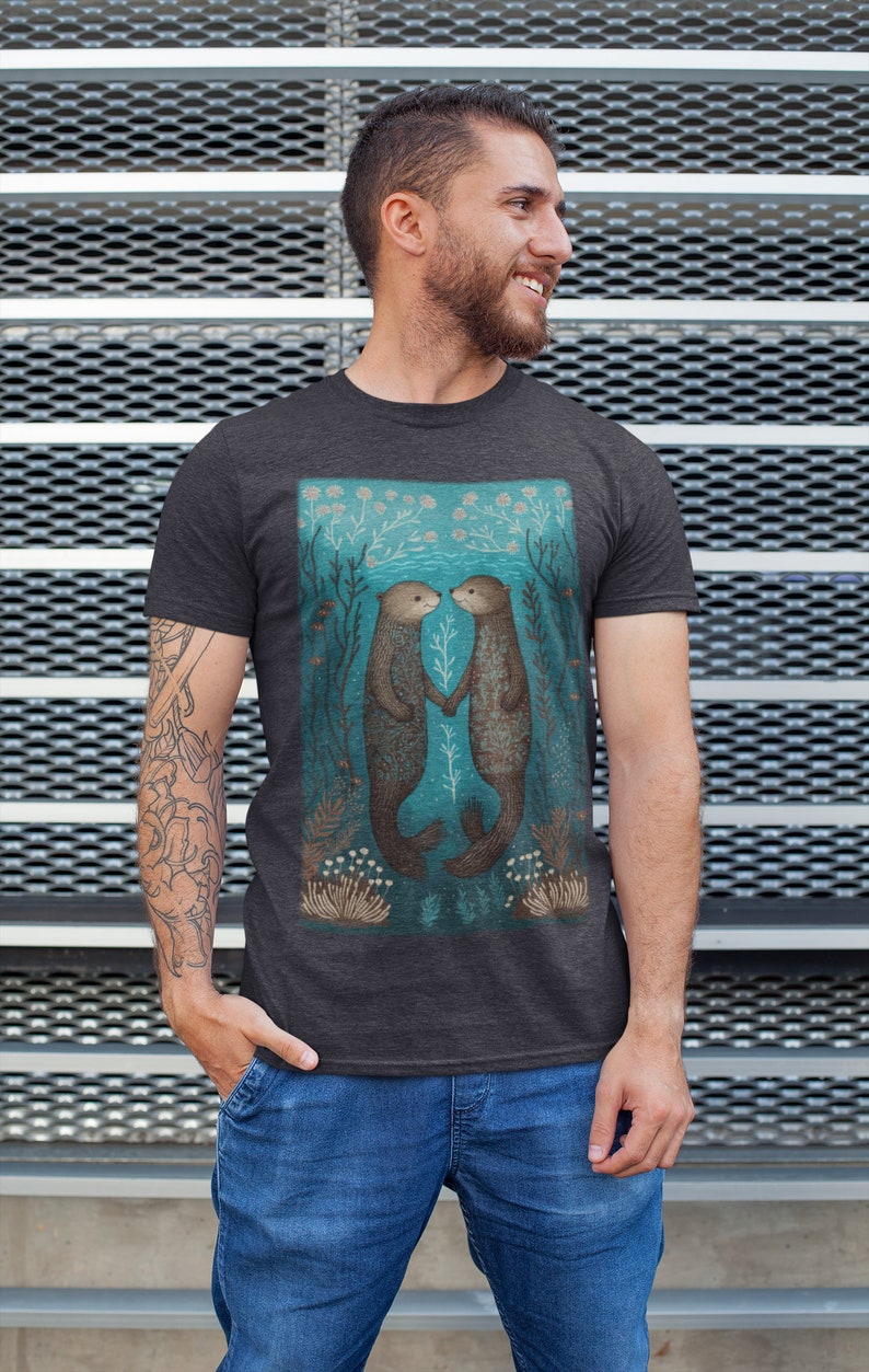 May include: A dark heather grey t-shirt featuring a graphic of two sea otters holding hands in a turquoise underwater scene. The otters are brown with white markings. The scene is surrounded by green and white seaweed and coral.