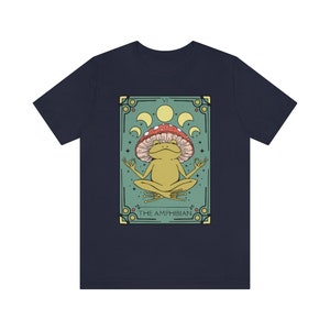 May include: A navy blue t-shirt with a graphic of a green frog sitting in a meditative pose on a mushroom. The graphic is surrounded by a green and gold frame with the text "VI THE AMPHIBIAN".