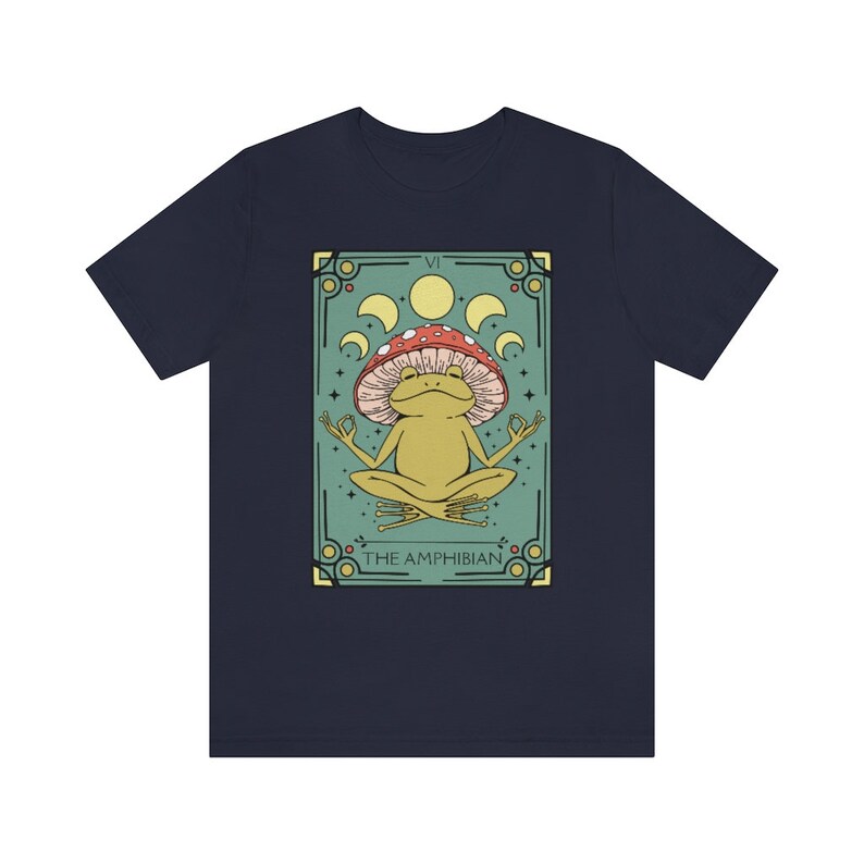May include: A navy blue t-shirt with a graphic of a green frog sitting in a meditative pose on a mushroom. The graphic is surrounded by a green and gold frame with the text "VI THE AMPHIBIAN".