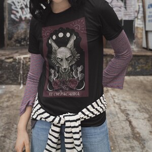 May include: Black t-shirt with a graphic print of a wolf-like creature with the text "El Chupacabra" underneath. The graphic is surrounded by a red border and features a crescent moon and stars.