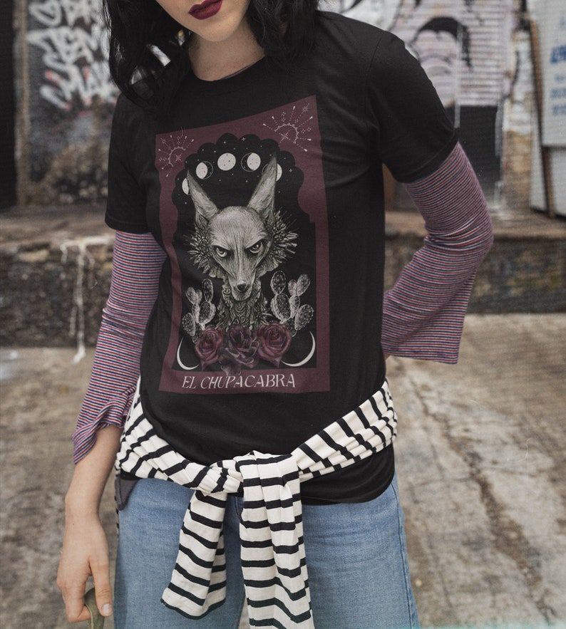 May include: Black t-shirt with a graphic print of a wolf-like creature with the text "El Chupacabra" underneath. The graphic is surrounded by a red border and features a crescent moon and stars.