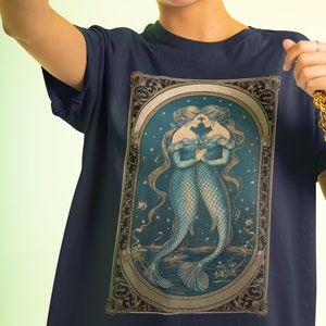 Puede incluir: Una camiseta azul marino con una ilustraci&oacute;n de estilo vintage de dos sirenas con cabello y colas flotantes, sobre un fondo de cielo estrellado. La ilustraci&oacute;n est&aacute; enmarcada con un borde dorado adornado.
