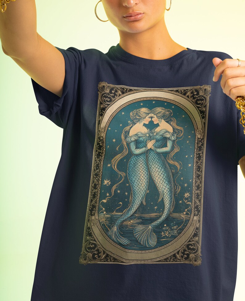 Puede incluir: Una camiseta azul marino con una ilustraci&oacute;n de estilo vintage de dos sirenas con cabello y colas flotantes, sobre un fondo de cielo estrellado. La ilustraci&oacute;n est&aacute; enmarcada con un borde dorado adornado.