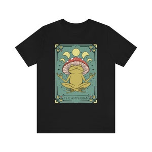 May include: A black t-shirt with a green and gold illustration of a frog wearing a red and white mushroom on its head. The illustration is surrounded by a decorative frame with the text "VI THE AMPHIBIAN".