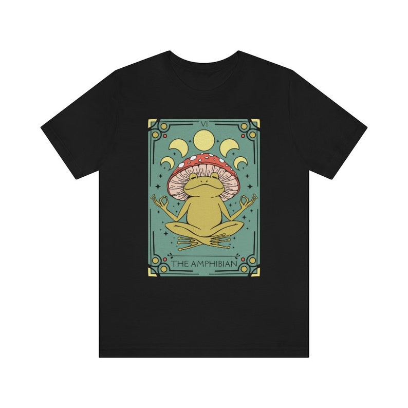 May include: A black t-shirt with a green and gold illustration of a frog wearing a red and white mushroom on its head. The illustration is surrounded by a decorative frame with the text "VI THE AMPHIBIAN".