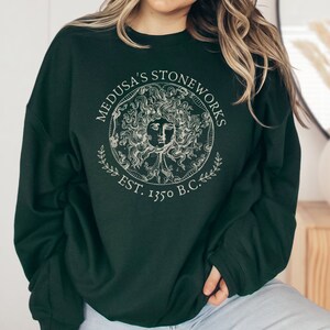 Peut inclure: Un sweat-shirt vert fonc&eacute; avec un imprim&eacute; graphique blanc de Medusa's Stoneworks, repr&eacute;sentant une image stylis&eacute;e de la t&ecirc;te de M&eacute;duse avec des serpents pour cheveux, entour&eacute;e d'un cercle avec le texte "Medusa's Stoneworks Est. 1350 av. J.-C."