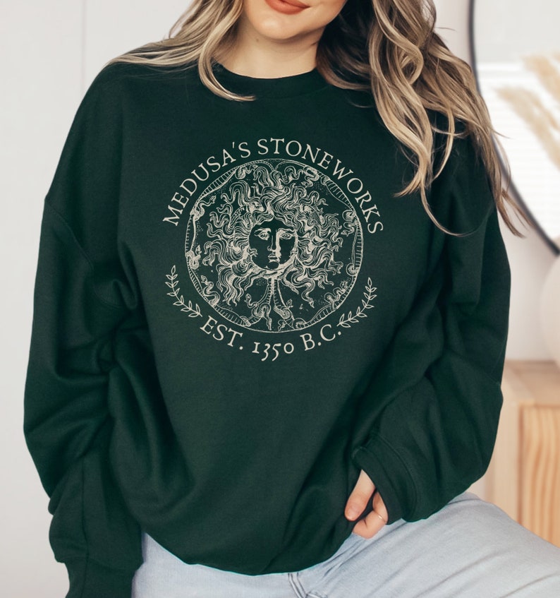 Peut inclure: Un sweat-shirt vert fonc&eacute; avec un imprim&eacute; graphique blanc de Medusa's Stoneworks, repr&eacute;sentant une image stylis&eacute;e de la t&ecirc;te de M&eacute;duse avec des serpents pour cheveux, entour&eacute;e d'un cercle avec le texte "Medusa's Stoneworks Est. 1350 av. J.-C."