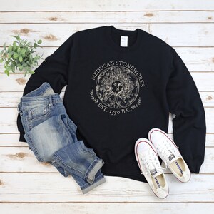 Peut inclure: Sweat-shirt noir avec un motif dor&eacute; de Medusa's Stoneworks, fond&eacute; en 1350 avant J.-C., sur un fond en bois blanc. Le sweat-shirt est associ&eacute; &agrave; un jean bleu et des baskets blanches.