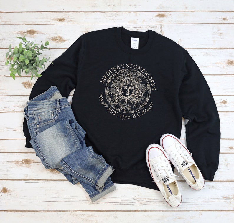 Peut inclure: Sweat-shirt noir avec un motif dor&eacute; de Medusa's Stoneworks, fond&eacute; en 1350 avant J.-C., sur un fond en bois blanc. Le sweat-shirt est associ&eacute; &agrave; un jean bleu et des baskets blanches.