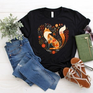 May include: Black t-shirt with a floral design featuring a fox in the center. The fox is orange, brown, and white with a floral pattern. The design is surrounded by green leaves and flowers.