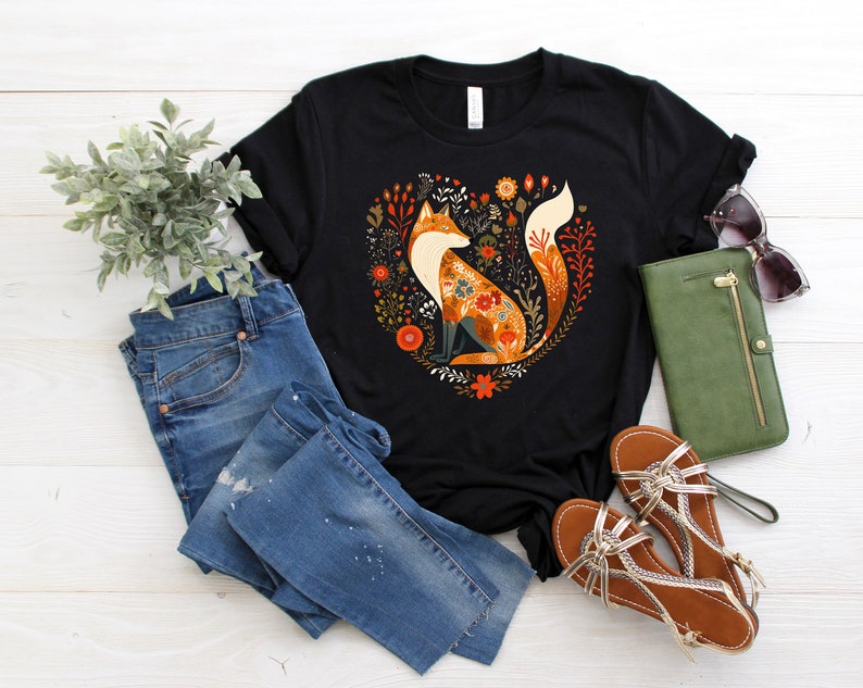 May include: Black t-shirt with a floral design featuring a fox in the center. The fox is orange, brown, and white with a floral pattern. The design is surrounded by green leaves and flowers.