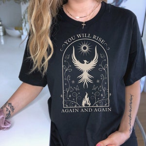 May include: Black t-shirt with a gold graphic of a phoenix rising from flames. The graphic is surrounded by a floral design and the text "You Will Rise Again and Again".
