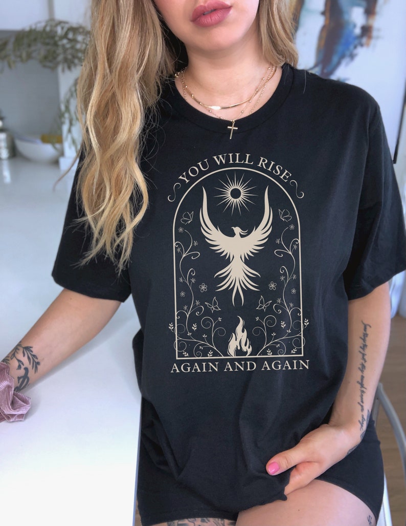 May include: Black t-shirt with a gold graphic of a phoenix rising from flames. The graphic is surrounded by a floral design and the text "You Will Rise Again and Again".
