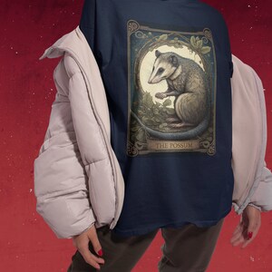 May include: A navy blue t-shirt with a vintage-style illustration of a possum. The illustration is surrounded by a gold frame and has the text "THE POSSUM" at the bottom.