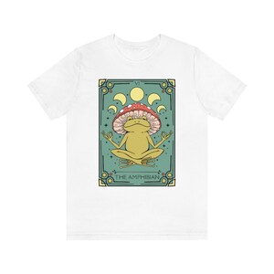 May include: White t-shirt with a green and gold illustration of a frog wearing a red and white mushroom on its head. The frog is sitting in a meditative pose with its eyes closed. The illustration is surrounded by a gold frame and the text "THE AMPHIBIAN" is printed below the frog.