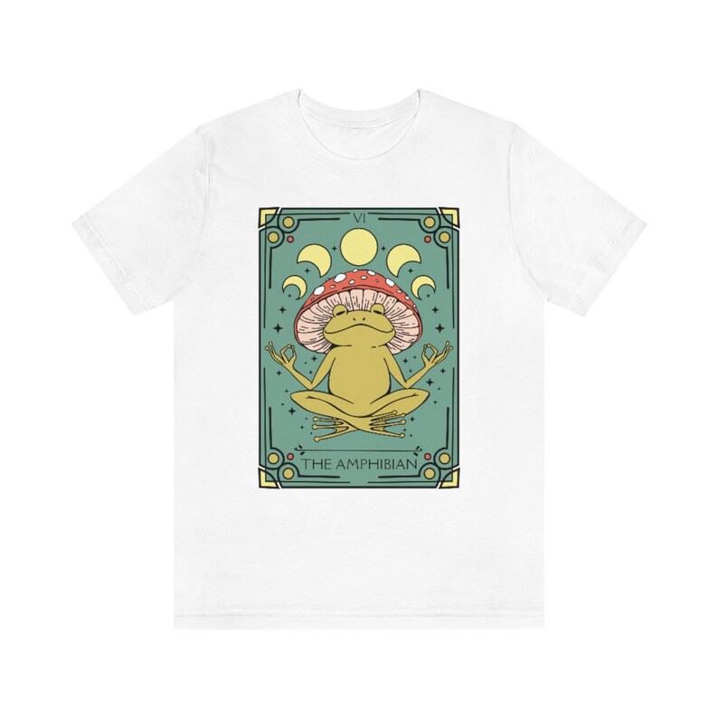 May include: White t-shirt with a green and gold illustration of a frog wearing a red and white mushroom on its head. The frog is sitting in a meditative pose with its eyes closed. The illustration is surrounded by a gold frame and the text "THE AMPHIBIAN" is printed below the frog.