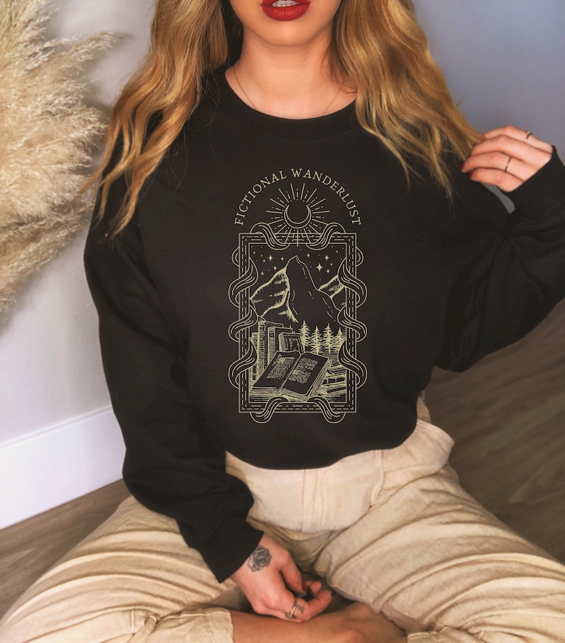 Op de afbeelding: Zwart sweatshirt met een gouden grafisch ontwerp met een bergketen, een wassende maan, sterren en een stapel boeken. De tekst "Fictional Wanderlust" staat in een cirkelvormig frame rond het ontwerp.