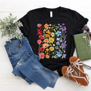 Puede incluir: Una camiseta negra con estampado floral arco&iacute;ris. La camiseta se combina con jeans azules, sandalias y una cartera verde.