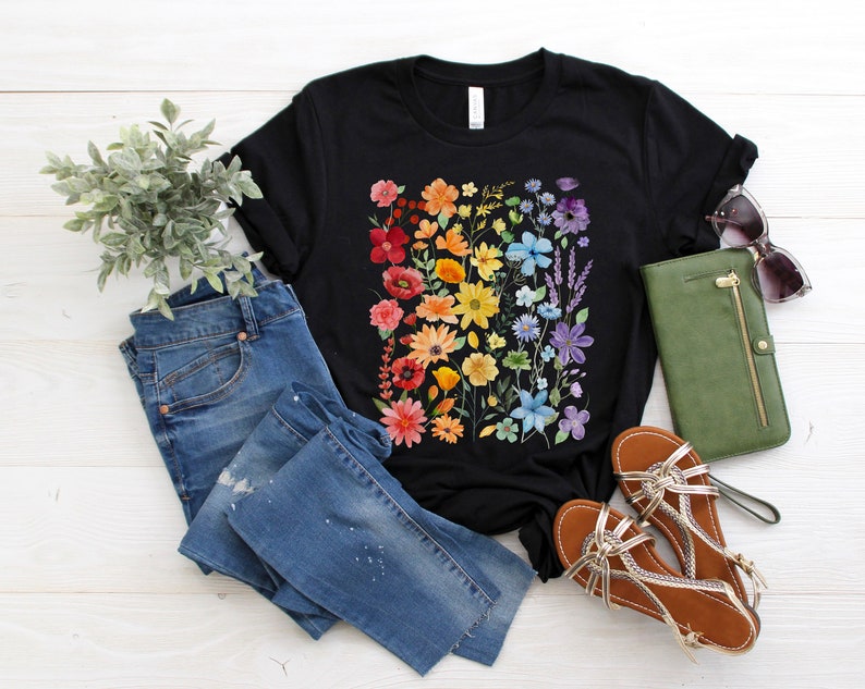 Puede incluir: Una camiseta negra con estampado floral arco&iacute;ris. La camiseta se combina con jeans azules, sandalias y una cartera verde.