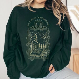 Op de afbeelding: Een donkergroen sweatshirt met een gouden grafische print van een bergketen, een wassende maan, sterren en boeken. De tekst "Fictional Wanderlust" is in een gouden lettertype rond de afbeelding gedrukt.