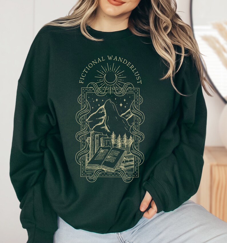 Op de afbeelding: Een donkergroen sweatshirt met een gouden grafische print van een bergketen, een wassende maan, sterren en boeken. De tekst "Fictional Wanderlust" is in een gouden lettertype rond de afbeelding gedrukt.