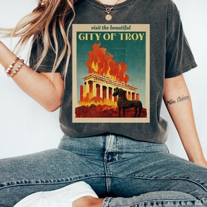 May include: A dark gray t-shirt with a vintage travel poster design featuring a burning building with a horse in front of it. The text on the poster reads "visit the beautiful CITY OF TROY".