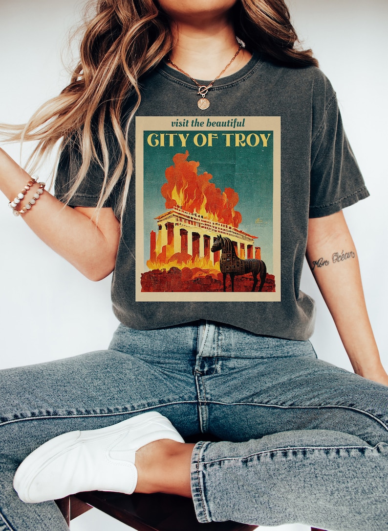 May include: A dark gray t-shirt with a vintage travel poster design featuring a burning building with a horse in front of it. The text on the poster reads "visit the beautiful CITY OF TROY".