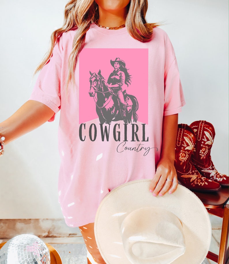 May include: Pink t-shirt with a graphic of a cowgirl riding a horse. The graphic is on a pink background and the text "COWGIRL Country" is below the graphic.