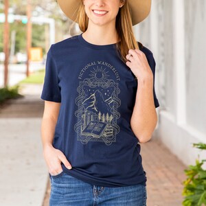 K&ouml;nnte beinhalten: Ein marineblaues T-Shirt mit einem goldenen Grafikdesign, das eine Bergkette, ein Buch und den Text "Fictional Wanderlust" zeigt.