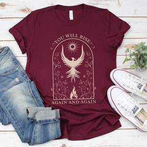 May include: A maroon t-shirt with a white graphic of a phoenix rising from flames. The graphic is surrounded by a floral design and the text "You Will Rise Again and Again".