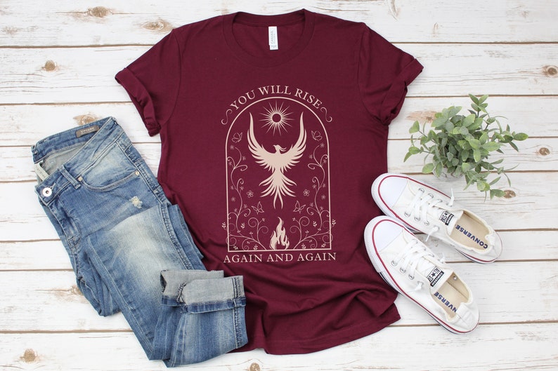May include: A maroon t-shirt with a white graphic of a phoenix rising from flames. The graphic is surrounded by a floral design and the text "You Will Rise Again and Again".