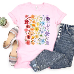 Puede incluir: Una camiseta rosa con estampado floral arco&iacute;ris. La camiseta se combina con jeans azules y pisos estampados.