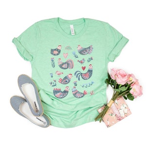K&ouml;nnte beinhalten: Ein hellgr&uuml;nes T-Shirt mit einer bunten Illustration von H&uuml;hnern und Blumen. Das Shirt ist aus einem weichen, bequemen Material.