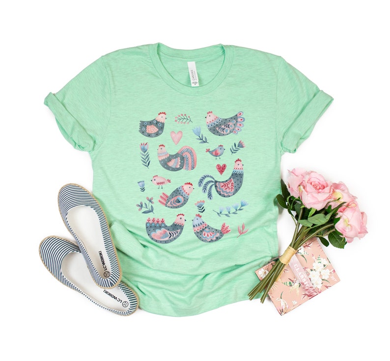 K&ouml;nnte beinhalten: Ein hellgr&uuml;nes T-Shirt mit einer bunten Illustration von H&uuml;hnern und Blumen. Das Shirt ist aus einem weichen, bequemen Material.