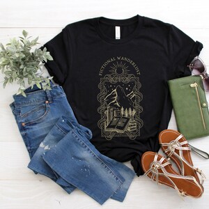 K&ouml;nnte beinhalten: Schwarzes T-Shirt mit einem goldenen Grafikdesign, das einen Berg, ein Buch und den Text "Fictional Wandererlust" zeigt.