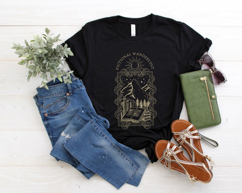K&ouml;nnte beinhalten: Schwarzes T-Shirt mit einem goldenen Grafikdesign, das einen Berg, ein Buch und den Text "Fictional Wandererlust" zeigt.