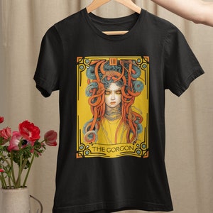 以下が含まれることがあります： 蛇の髪をした女性のグラフィックプリントが施された黒いTシャツ。グラフィックは黄色、オレンジ、青の色合いで、下部に「THE GORGON」の文字があります。