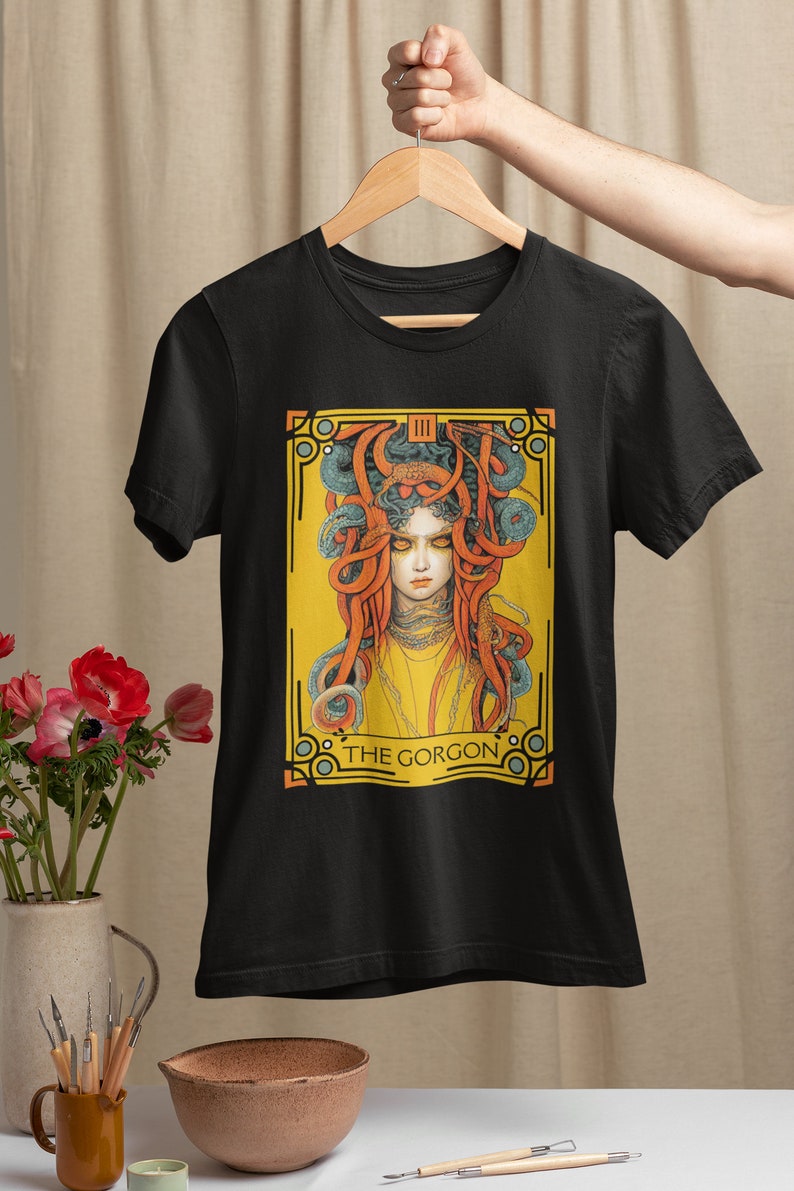 以下が含まれることがあります： 蛇の髪をした女性のグラフィックプリントが施された黒いTシャツ。グラフィックは黄色、オレンジ、青の色合いで、下部に「THE GORGON」の文字があります。