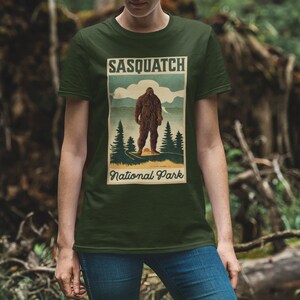 May include: Dark green t-shirt featuring a vintage-style graphic of a Sasquatch in a forest setting. The graphic includes the text "SASQUATCH National Park" in a retro font. The shirt is made of a soft material.