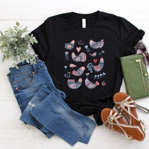 K&ouml;nnte beinhalten: Schwarzes T-Shirt mit einem bunten Muster aus stilisierten V&ouml;geln und Herzen. Das Shirt liegt auf einer wei&szlig;en Holzoberfl&auml;che mit einer blauen Jeans, einer gr&uuml;nen Geldb&ouml;rse und braunen Sandalen.