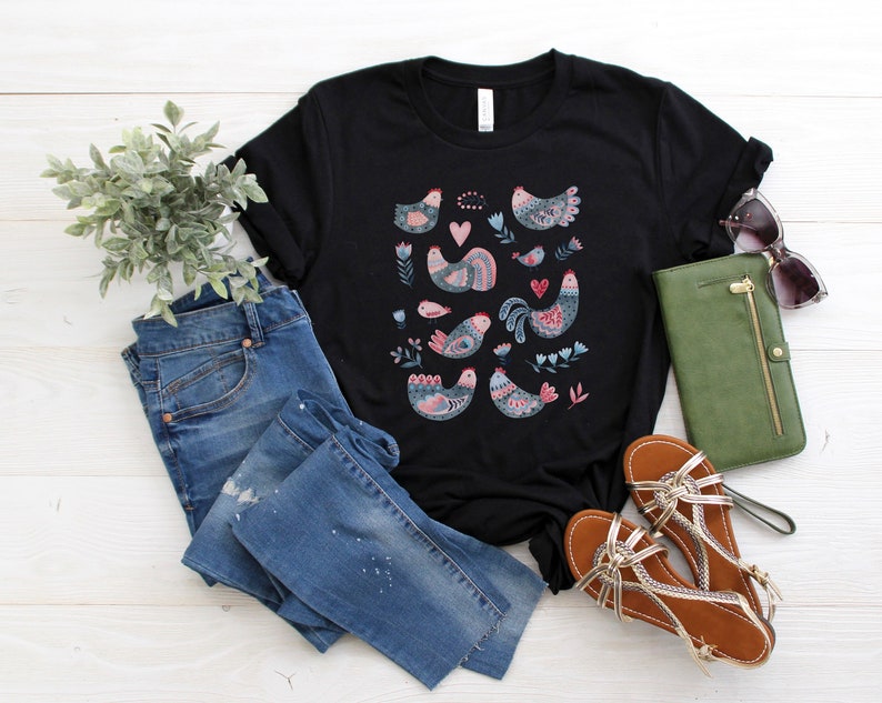 K&ouml;nnte beinhalten: Schwarzes T-Shirt mit einem bunten Muster aus stilisierten V&ouml;geln und Herzen. Das Shirt liegt auf einer wei&szlig;en Holzoberfl&auml;che mit einer blauen Jeans, einer gr&uuml;nen Geldb&ouml;rse und braunen Sandalen.