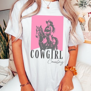 May include: White t-shirt with a pink graphic of a cowgirl riding a horse. The text "COWGIRL Country" is printed below the graphic.