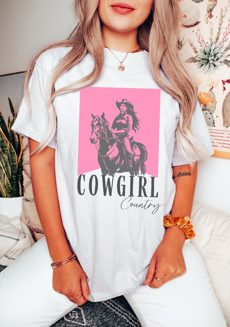 May include: White t-shirt with a pink graphic of a cowgirl riding a horse. The text "COWGIRL Country" is printed below the graphic.