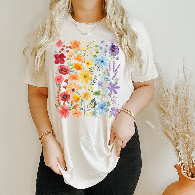 Puede incluir: Una camiseta blanca con un estampado floral arco&iacute;ris. Las flores est&aacute;n dispuestas en un patr&oacute;n arco&iacute;ris, con flores rojas, naranjas, amarillas, verdes, azules, &iacute;ndigo y violetas.