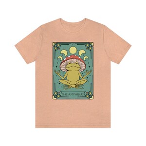 May include: A light peach t-shirt with a graphic of a green frog meditating under a red and white mushroom. The graphic is surrounded by a teal border and includes the text "VI THE AMPHIBIAN".