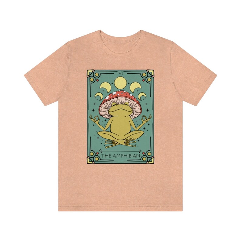 May include: A light peach t-shirt with a graphic of a green frog meditating under a red and white mushroom. The graphic is surrounded by a teal border and includes the text "VI THE AMPHIBIAN".