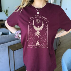 May include: A maroon t-shirt with a graphic of a phoenix rising from flames in a floral arch. The text "You Will Rise Again and Again" is printed below the graphic.